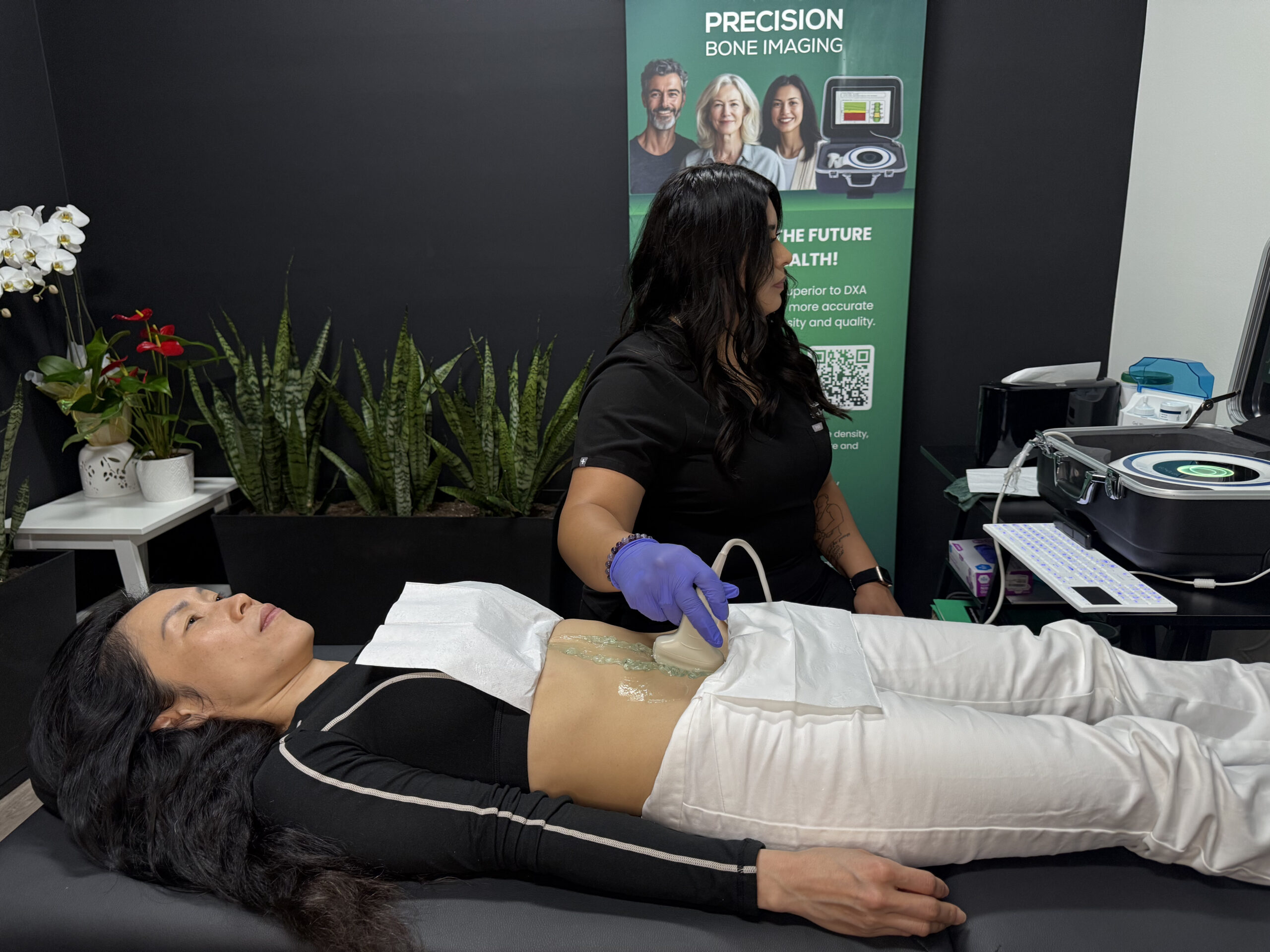 Technician wearing gloves performs a bone density scan on a woman lying on her back on an examination bed; the patient is wearing black and white clothing, and a Precision Bone Imaging banner and scanning equipment are visible.
