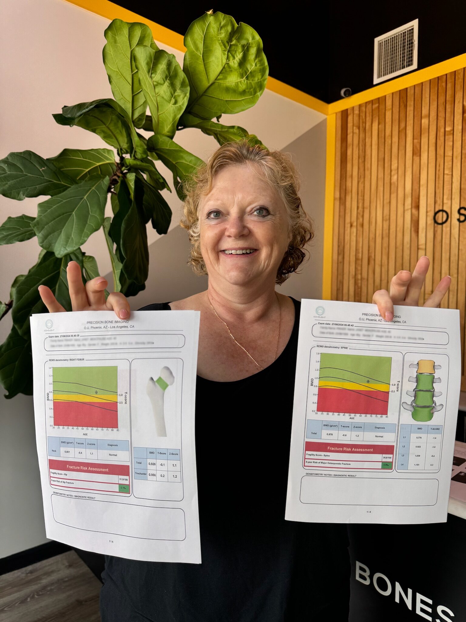 Smiling woman holding two bone density scan reports in front of a plant and wooden wall.